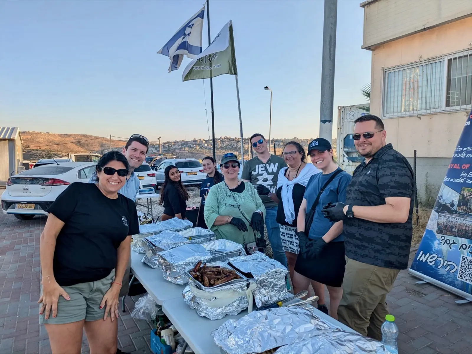 Atlanta Israel Coalition volunteers gathered with trays of food at an outdoor barbecue for Israeli soldiers, an Israeli flag flying overhead in the evening light.
