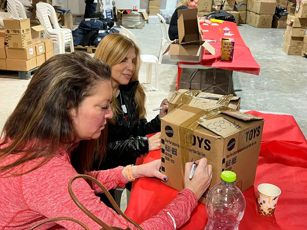 Two AIC volunteers write on taped-up boxes of toys at a long red table in a warehouse.
