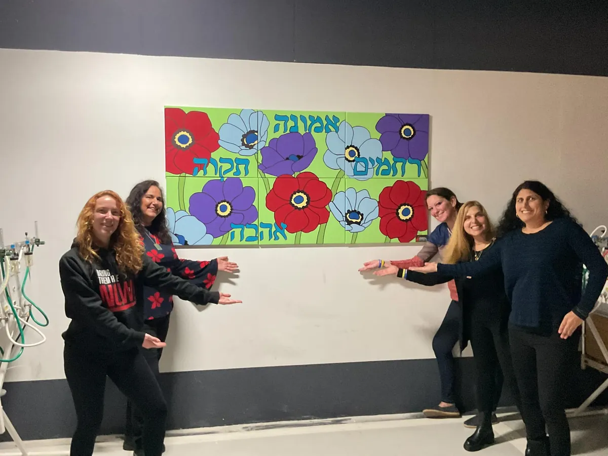 Five AIC volunteers point to a painted mural of red, blue and purple anemones with Hebrew words for faith, hope, love and compassion, hung on a hospital wall.