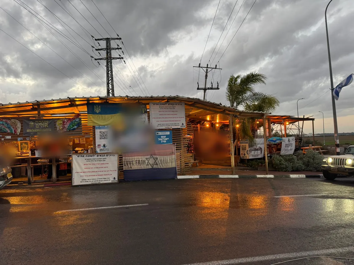 The Achim Shuva canteen at the junction at dusk, lit up under a stormy sky, an Israeli flag on the lamp post.