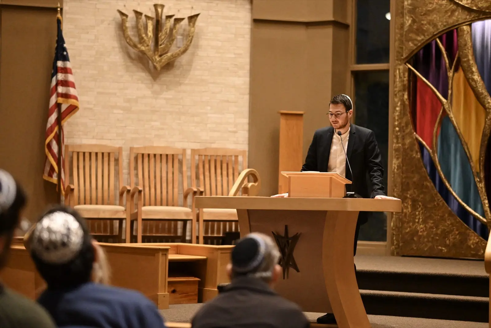 A speaker addresses a community audience from the bimah at a synagogue event, an American flag and menorah behind them.