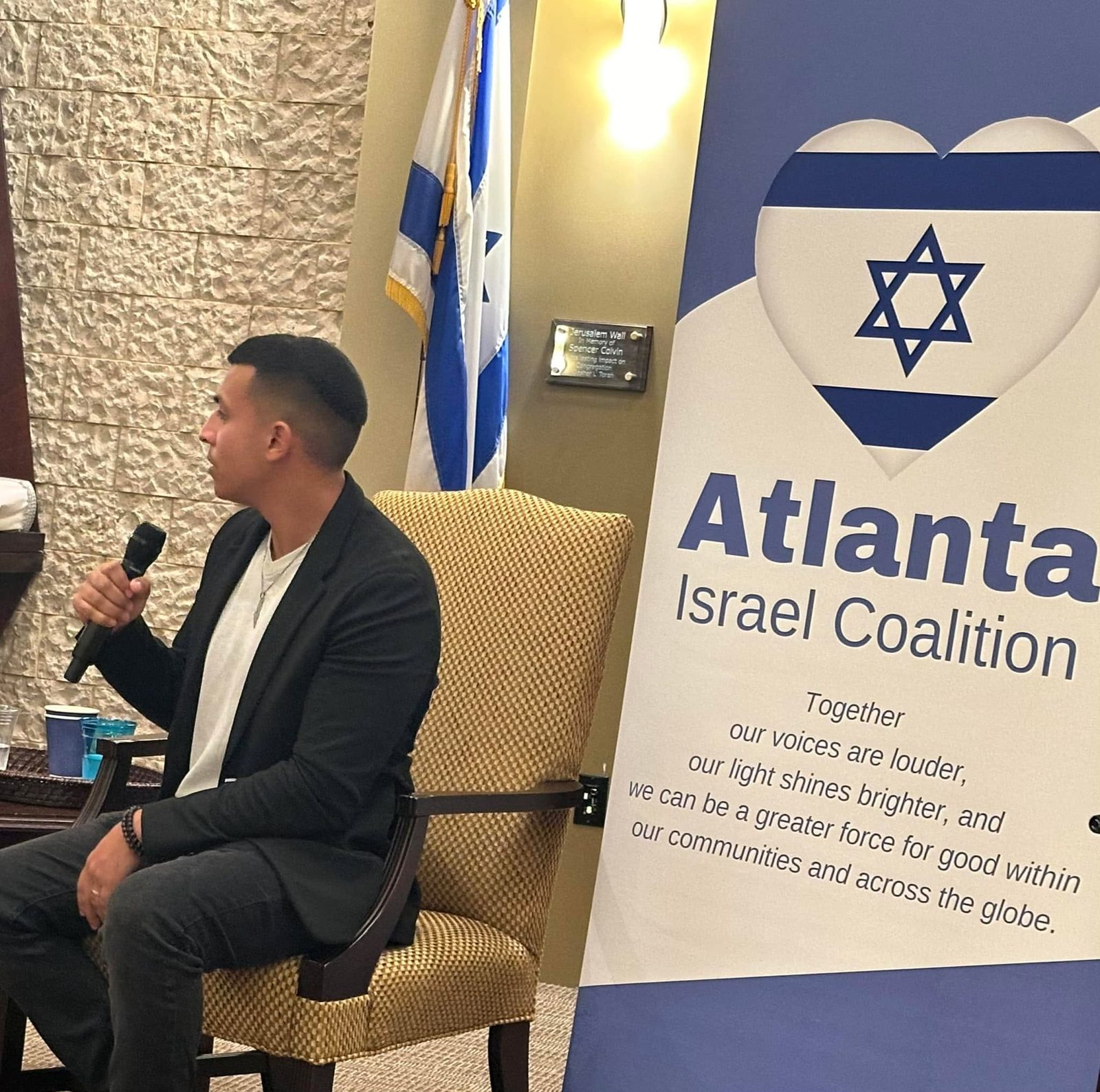 Yirmiyahu Danzig speaking into a microphone beside the Atlanta Israel Coalition banner at his evening talk.