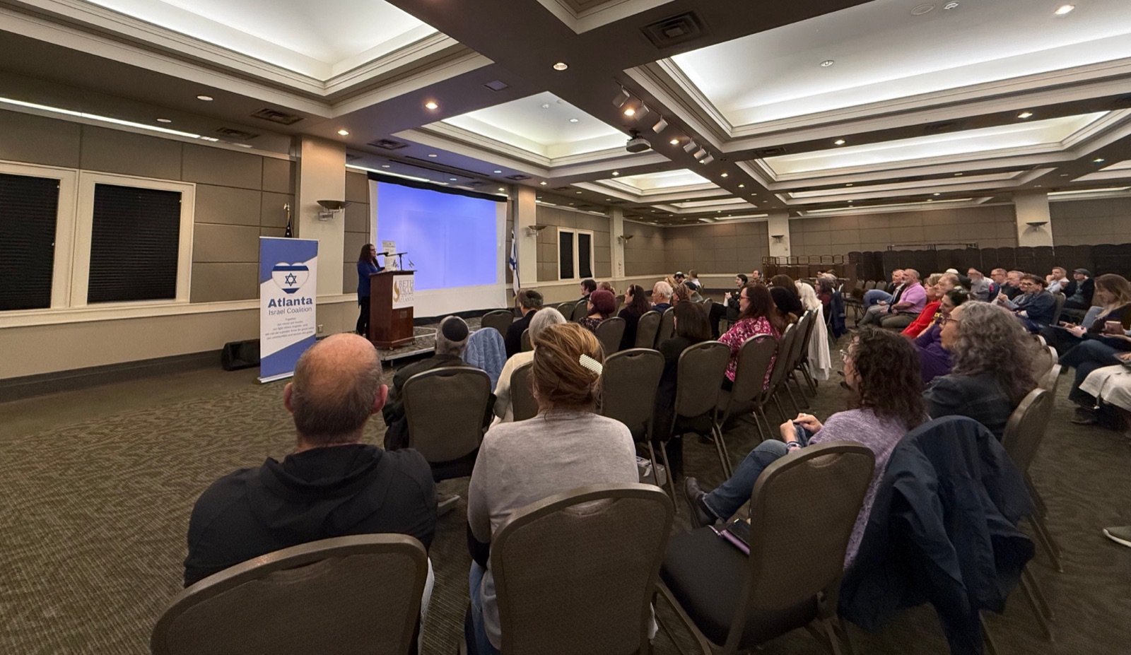 An Atlanta Israel Coalition speaker addresses a seated audience at a community briefing, an Israeli flag projected on the screen.