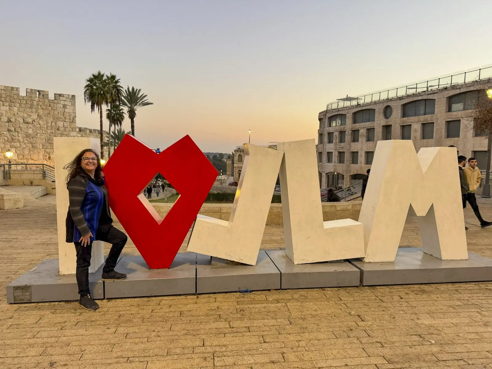 A visitor stands beside a large 'I love JLM' sculpture at sunset, the walls of Jerusalem’s Old City behind.