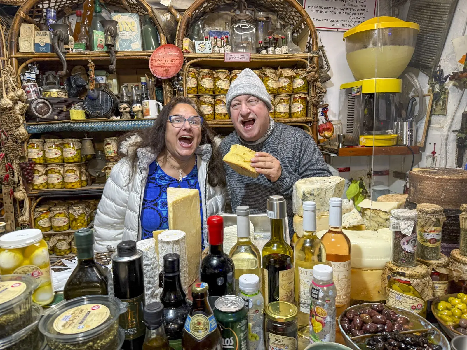 Two AIC visitors laugh together at a colorful Israeli market stall stacked with cheeses, olives, and preserves.