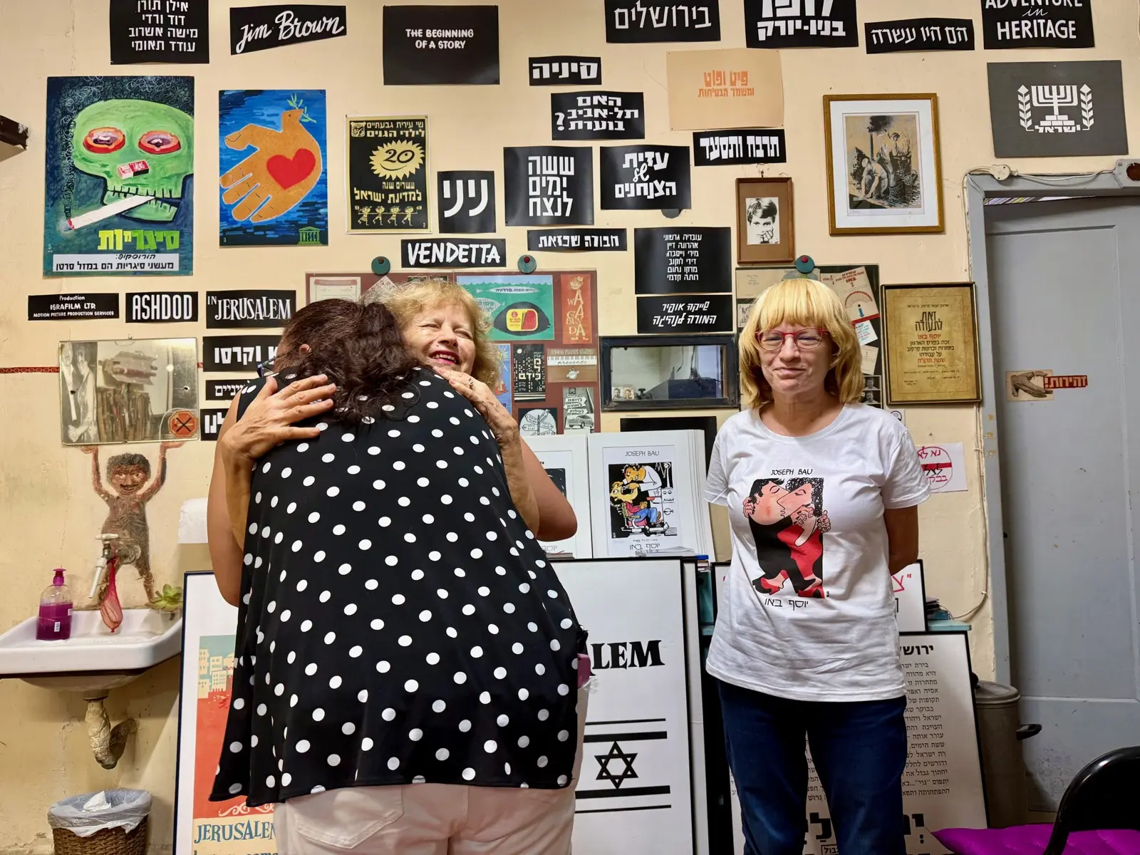 Two people embrace warmly during an Atlanta Israel Coalition visit, in a room lined with Hebrew posters and Israeli artwork.