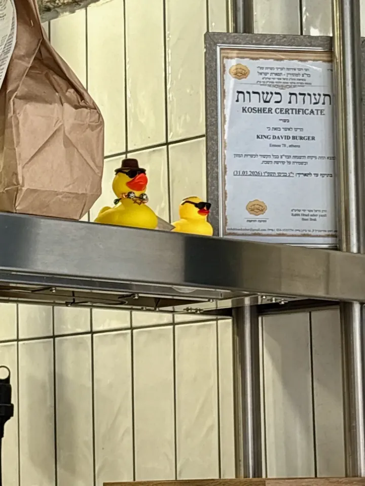 Two yellow kosher ducks in sunglasses on a restaurant shelf in Athens, beside a framed kosher certificate.